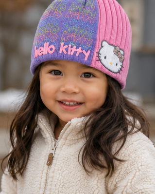 A young child wearing a pink and purple Hello Kitty ribbed knit beanie hat and a cream fleece zip jacket, photographed outdoors with a blurred background.