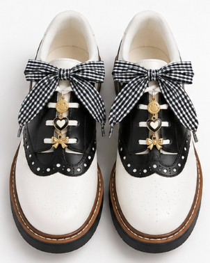 A pair of black and white leather lace-up shoes with black-and-white gingham ribbon bows, gold heart-shaped laces, and decorative gold charms, shown on a white background.