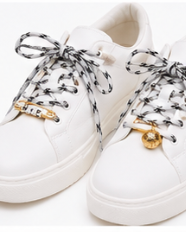 A pair of white leather low-top sneakers with black and white patterned laces, featuring gold-tone metal eyelets and a gold circular charm on the laces, set against a plain white background.