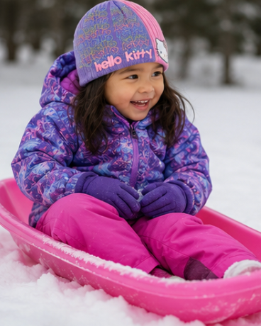 A young child sitting in a pink plastic sled on snow, wearing a purple and pink winter jacket, bright pink pants, and a pink and purple Hello Kitty beanie.