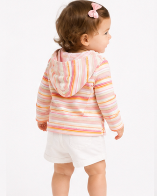 A toddler girl seen from the back wearing a pastel striped hooded sweatshirt with white shorts and white sandals, standing against a plain white studio background.