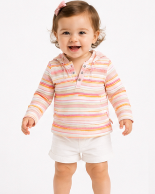 A toddler girl smiling and standing on a white studio background, wearing a pastel striped long-sleeve polo shirt with white shorts and white sandals.